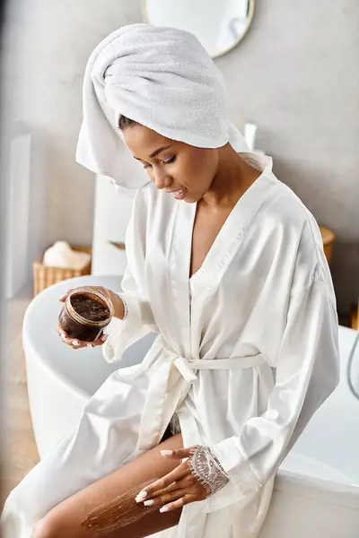 An African American woman in a bathrobe relaxes with a coffee scrub in her modern bathroom, embracing a moment of calm and beauty.