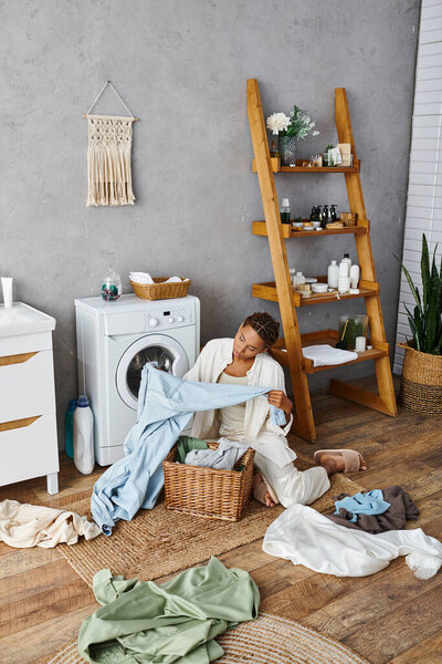A woman with afro braids sits next to a washing machine, doing laundry in a bathroom setting.