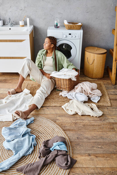 African American woman with afro braids sitting by washing machine in a bathroom, engaged in doing laundry.