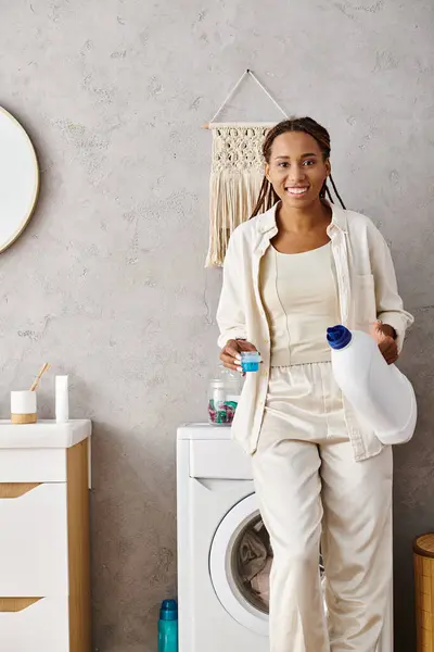 stock image An African American woman with afro braids stands next to a washing machine, doing laundry in a bathroom.