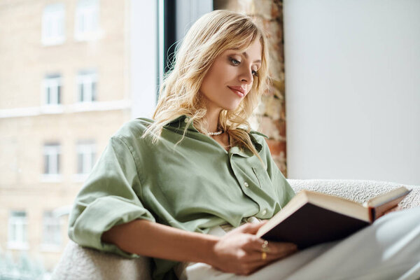 Woman in chair reading book in kitchen of apartment.