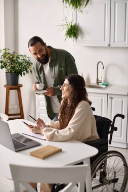 Yakışıklı adam, tekerlekli sandalyedeki engelli karısının yanında kahve içiyor ve laptopta çalışıyor.