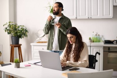 Yakışıklı adam, tekerlekli sandalyedeki engelli karısının yanında kahve içiyor ve laptopta çalışıyor.