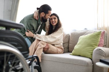 Ev kıyafetleri içinde sevgi dolu bir adam eşine tekerlekli sandalyeden kanepeye oturması için yardım ediyor.