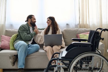 Keyifli bir adam evdeki kanepede otururken engelli, neşeli karısının yanında dizüstü bilgisayara bakıyor.