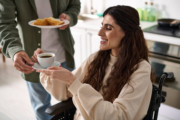 cheerful disabled woman in wheelchair holding coffee cup next to her husband while in kitchen