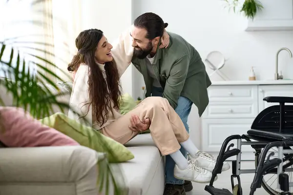車椅子からソファに取得する移動性障害を持つ彼の妻を助けるホームウェアで愛する男 — ストック写真