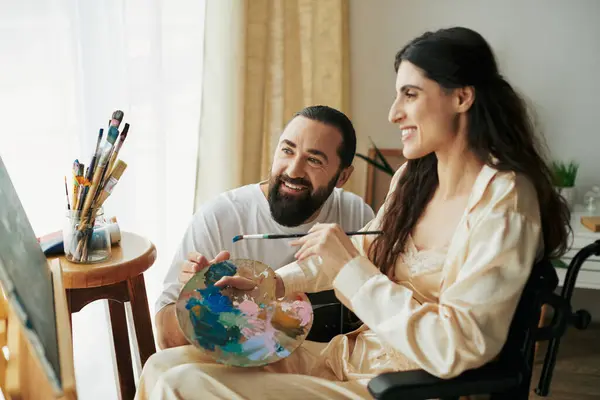 stock image positive husband helping his inclusive beautiful wife on wheelchair to paint on easel while at home