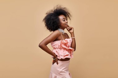 20 'li yaşlardaki mutlu Afro-Amerikalı kız şeftali rengi bluz ve pastel etekle poz veriyor.