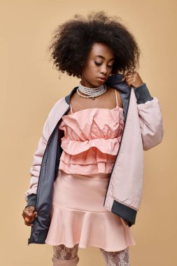20 'li yaşlarda Afro-Amerikan bir kız, Pastel kıyafetler giymiş, bej arka planda ceketi var, şeftali rengi tüylü.