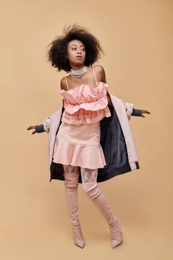 20 'li yaşlarda genç bir Afro-Amerikan kadın, şeftali rengi kıyafetler içinde, ceketi ve bej çizmeleriyle poz veriyor.