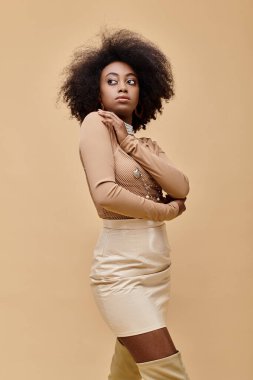 Pastel şeftali rengi elbisesiyle bej arka planda poz veren kıvırcık saçlı Afrikalı Amerikalı genç bir kız.
