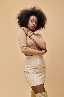 Pastel şeftali rengi elbisesiyle bej arka planda poz veren kıvırcık saçlı Afro-Amerikan model.