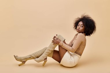 curly african american girl in trendy peach fuzz outfit and thigh-high boots reclines on a beige clipart