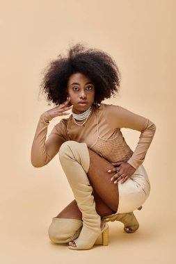 curly african american model in trendy peach fuzz outfit and thigh-high boots sitting on a beige clipart