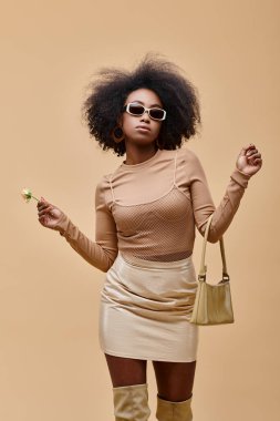 Güneş gözlüklü şık bir Afro-Amerikan modeli, el çantası ve bej arka planda küçük bir gül taşıyor.
