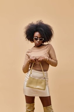 Genç ve şık Afro-Amerikalı güneş gözlüklü kadın bej arka planda modaya uygun el çantası taşıyor.
