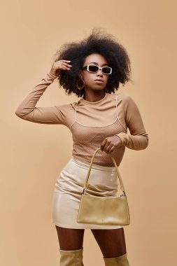 Güneş gözlüklü genç ve şık Afro-Amerikan modeli bej arka planda modaya uygun el çantası taşıyor.
