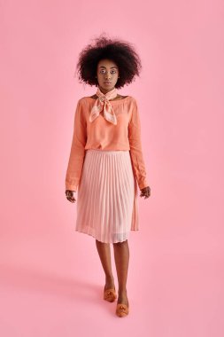 Şeftali rengi bluz ve pastel pembe arka planda poz veren midi etekli zarif Afrikalı Amerikalı kadın.