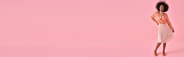 Şeftali rengi bluz ve midi etekli zarif Afro-Amerikan kadın pastel pembe arka planda poz veriyor.
