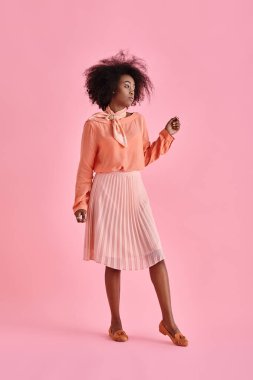 Şeftali rengi bluz ve pastel pembe arka planda poz veren midi etekli rüya gibi Afrikalı Amerikalı kadın.