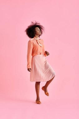 Şeftali rengi bluzlu kıvırcık Afrikalı Amerikalı kadın ve pastel pembe arka planda poz veren midi etekli.
