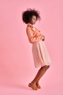 Şeftali rengi bluz ve midi etekli kıvırcık Afrikalı Amerikalı kadın pastel pembe arka plana bakıyor.