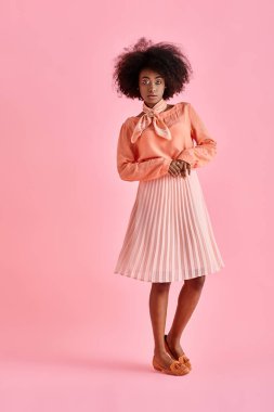 Şeftali rengi bluz ve midi etekli kıvırcık Afrikalı Amerikalı kadın pastel pembe arka planda poz veriyor.