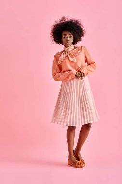 curly african american woman in peach blouse and midi skirt strikes a pose on pastel pink background clipart