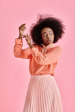 Şeftali rengi bluz, etek ve boyun atkısı giymiş, pastel pembe arka planda poz veren genç Afrikalı Amerikalı kadın.