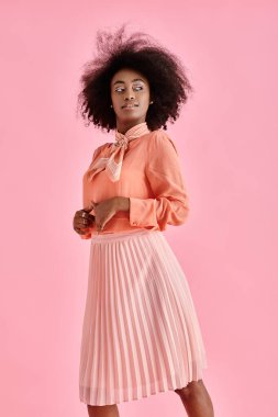 Şeftali rengi bluz, etek ve boyun atkısı giymiş, pastel pembe arka planda poz veren kıvırcık Afrikalı Amerikalı kadın.