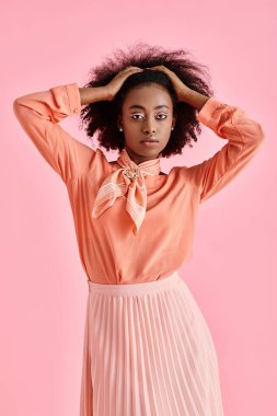 Şeftali rengi bluzlu Afro-Amerikan kadın, midi etek ve boyun eşarbı, pembe arka planda saçları düzeltiyor.