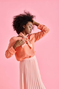 african american woman in peach blouse, midi skirt and scarf adjusting curly hair on pink backdrop clipart