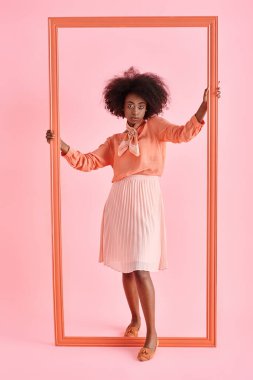 young african american woman in peach blouse and midi skirt strikes pose near frame on pink backdrop clipart