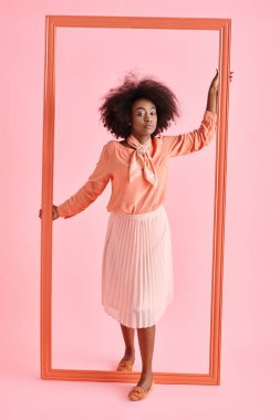 pretty african american woman in peach blouse and midi skirt strikes pose near frame on pastel pink clipart