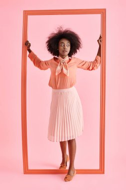Şeftali rengi bluz ve midi etekli genç Afrikalı Amerikalı kadın, pastel pembe çerçevenin yanında poz veriyor.