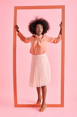young african american woman in peach blouse and midi skirt strikes pose near frame on pastel pink clipart
