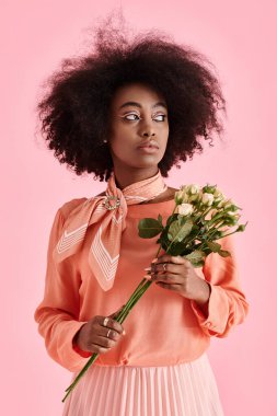 Şeftali tüyü bluzlu rüya gibi Afrikalı Amerikalı kız elinde çiçeklerle pembe arka plana bakıyor.
