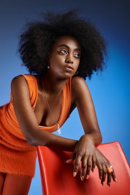 stylish african american woman in peach fuzz dress posing next to chair on vivid blue background clipart