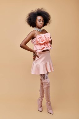 curly young african american woman in peach ruffled top and over knee boots on beige background clipart