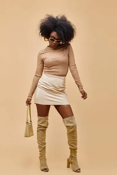 african american model in sunglasses and thigh-high boots holding trendy handbag on beige background