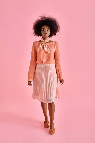 Graceful african american woman in peach blouse and midi skirt posing on pastel pink background