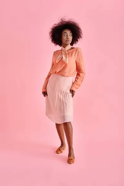 elegant african american woman in peach blouse and midi skirt posing on pastel pink background