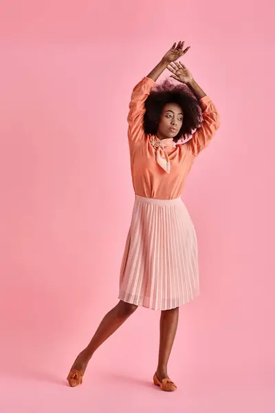 curly african american woman in peach blouse and midi skirt posing with raised hand on pastel pink
