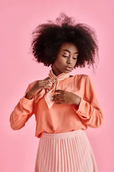 elegant african american woman in peach blouse adjusting neck scarf and posing on pink background