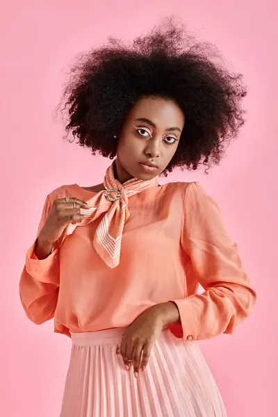 young african american woman in peach blouse adjusting neck scarf and posing on pink background