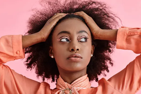portrait of african american woman in peach blouse and scarf adjusting curly hair on pink backdrop