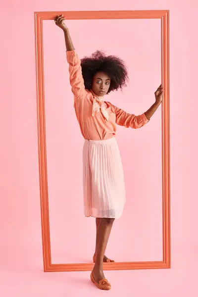 身穿桃色衬衫和迷你裙的年轻的非洲裔美国女人 在粉红的背景下摆出接近镜框的姿势 — 图库照片