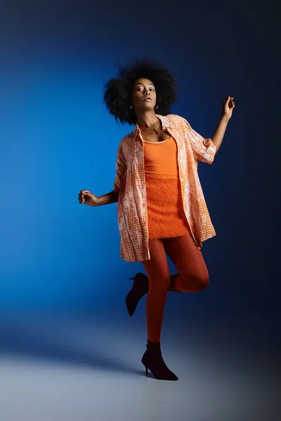 Chic look of african american model in patterned shirt and orange dress posing on blue background