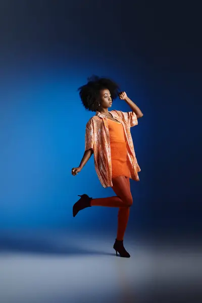 stylish look of african american woman in patterned shirt and orange dress posing on blue background